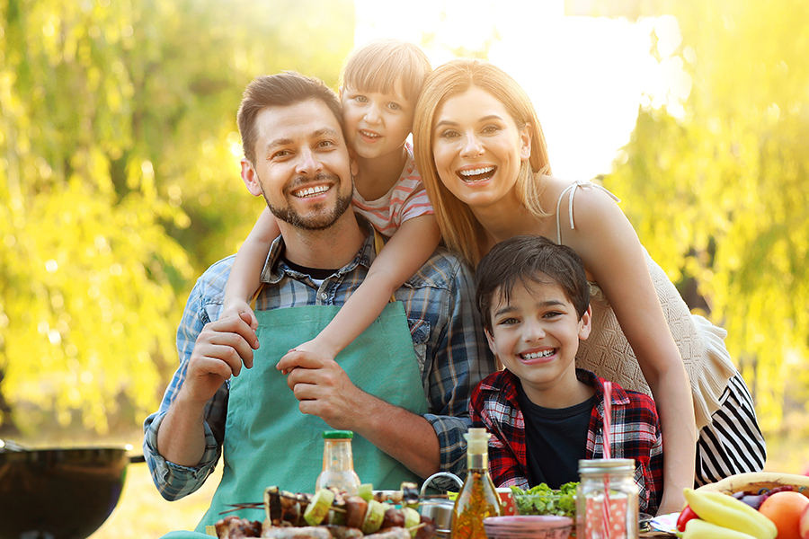 A cheerful family of four posing at a picnic table outdoors, surrounded by fresh vegetables and grilling supplies, bathed in warm sunlight and smiling brightly.