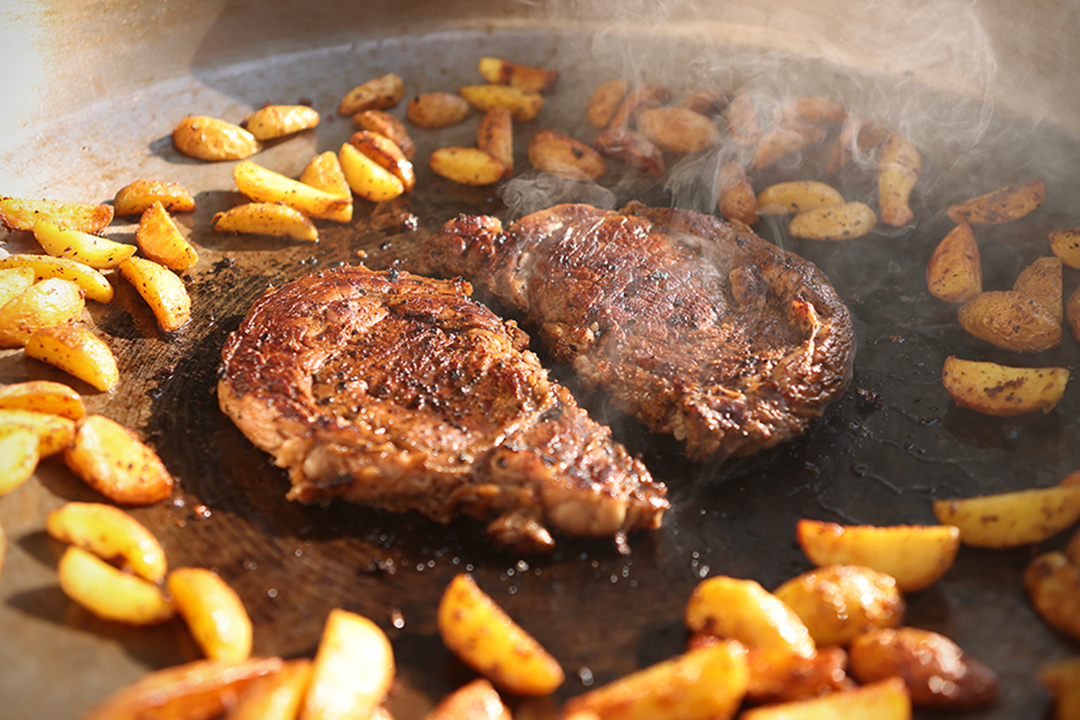 Two seared steaks sizzling on a flat-top grill surrounded by golden-brown potato wedges, showcasing a hearty Drummond Ranch meal in progress.