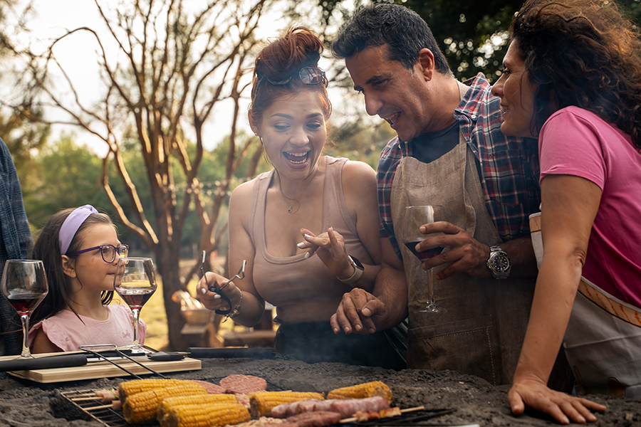 A lively outdoor cookout scene with adults and a child smiling and talking around a grill filled with corn, skewers, and burger patties, with glasses of wine adding to the festive mood.