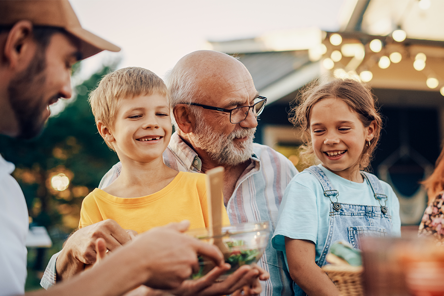 A grandfather surrounded by two smiling children and an adult as they prepare food outdoors, enjoying a multigenerational family gathering with warm, natural lighting.