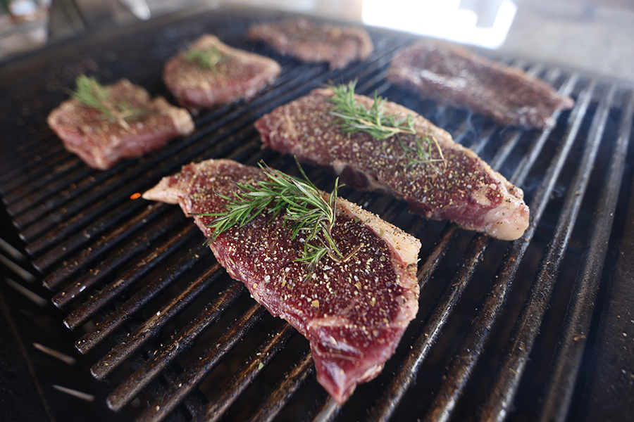 Several raw, seasoned steaks topped with fresh rosemary sprigs sizzling on a grill, with visible grill marks beginning to form as smoke rises in the background.